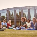 group of university or college students sit on grass outdoors on campus talking and working