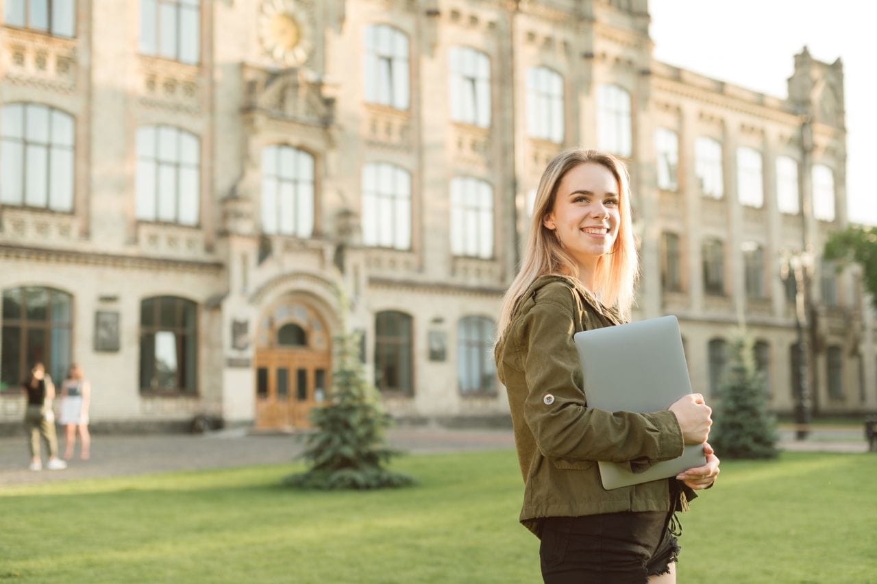 portrait of happy lady in casual clothes standing on grass in campus on background of university building with laptop in hands, looking away at copy space and smiling.girl is walking college yard