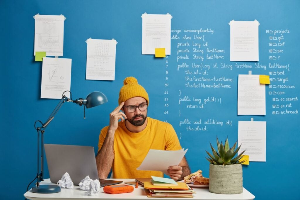 serious male employee or freelancer considers paper document, wears yellow hat and t shirt, studies on laptop computer online, works from home, looks through material, poses in coworking space